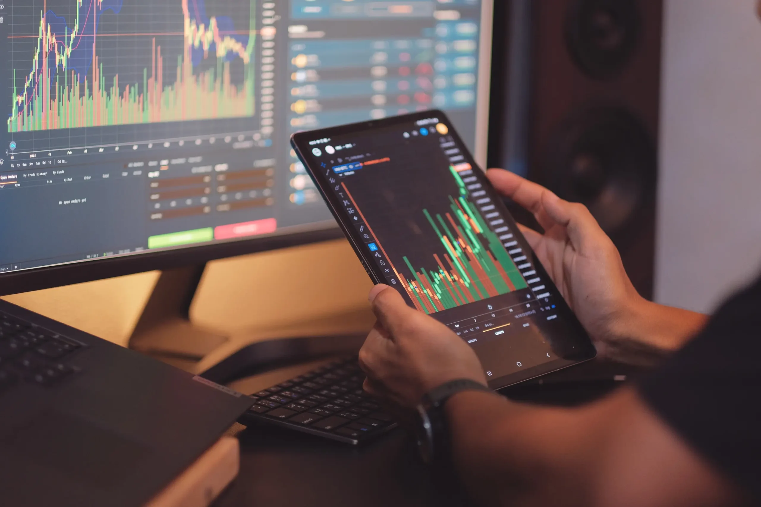 Investor reviewing equity candlestick charts on tablet while monitoring live markets on desktop screen.