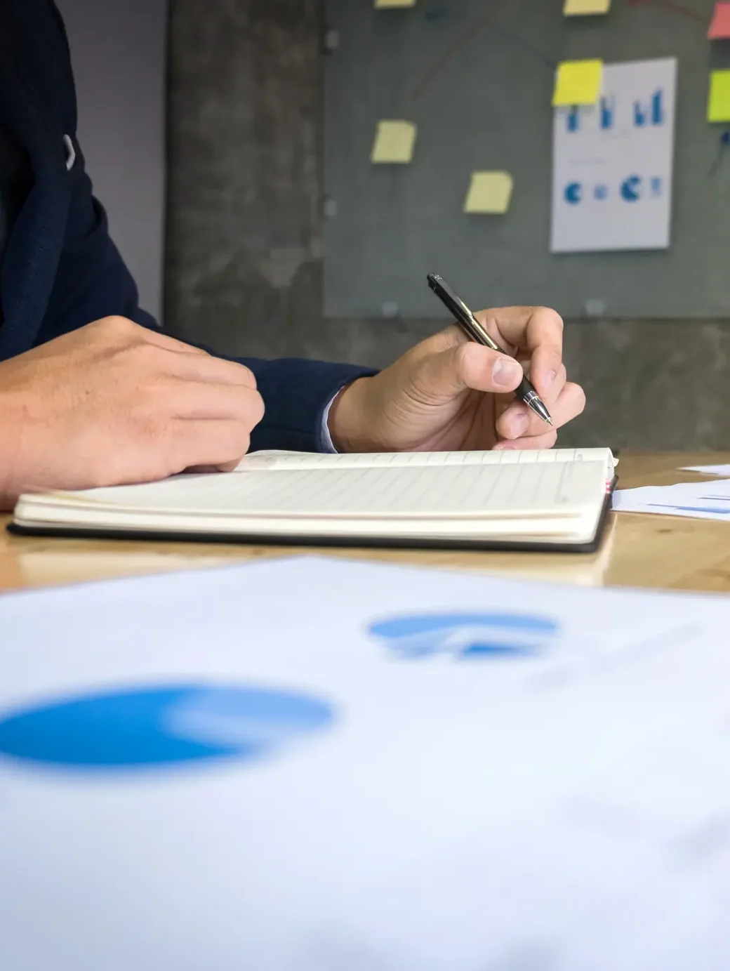 Investor taking notes while colleague presents portfolio performance charts on wall during strategy meeting.