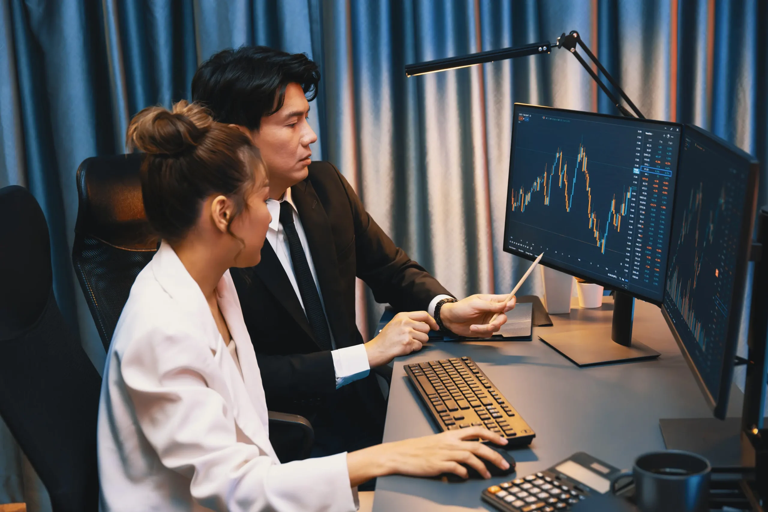 Two investment professionals reviewing live equity price charts on dual trading monitors in evening office.